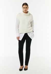 Kremowy sweter z golfem noszony na białej koszuli z guzikami, w połączeniu z czarnymi, dopasowanymi spodniami i czarnymi butami na obcasie. Prosty, teksturowany design.
