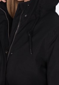 Zwarte jas met ritssluiting en drukknopen, voorzien van een capuchon en een trekkoord. Materiaal lijkt glad te zijn met een matte afwerking.