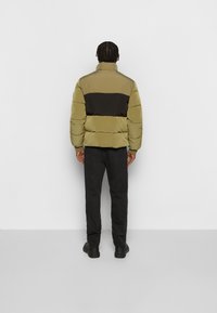 Puffjacka i olivgrön och svart. Har en hög krage och horisontell färgblockdesign. Bärs med svarta byxor och svarta skor.