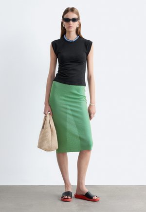 Femme porte un haut noir sans manches, une jupe midi rayée verte, des lunettes de soleil noires, un collier bleu, tient un sac beige tressé et des sandales slide noires et rouges.