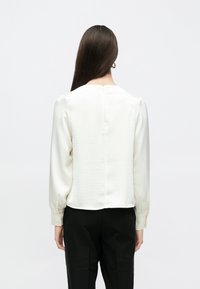 Hvid silke bluse med lange ærmer, rund halsudskæring, subtil glans og lukning bagpå. Serveret med sorte skræddersyede bukser.