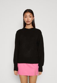 Schwarzer Pullover mit langen Ärmeln, runden Ausschnitt und strukturiertem Finish. Getragen mit einem pinken Minikleid mit seitlichem Schlitz.