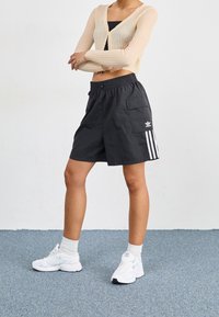 Pantaloni cargo neri con tasche laterali e tre strisce bianche sul lato sinistro. Abbinati a un cardigan a coste beige chiaro e sneakers bianche.
