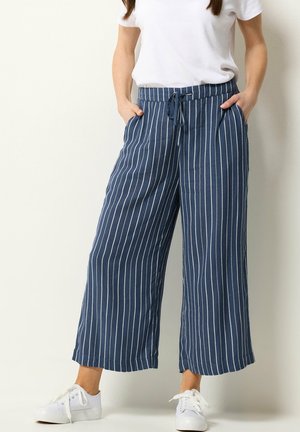 Frau trägt blaue, weit geschnittene Hose mit weißen vertikalen Streifen, weißes T-Shirt und weiße Sneakers, Hände in den Taschen, steht vor einer einfarbigen Wand.