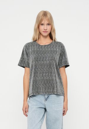 Ung kvinna med blont hår iklädd en kortärmad, grå texturerad topp och ljusblå jeans mot en vit bakgrund.