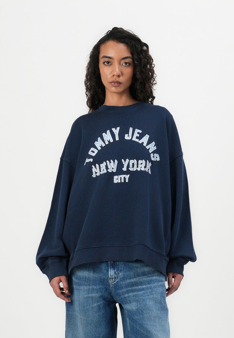Felpa oversize blu navy con grande scritta glitter bianca "TOMMY JEANS NEW YORK CITY." Tessuto morbido e polsini a costine.