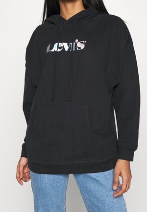 Person iført en sort Levi’s hættetrøje med metallisk logo, frontlomme og lyseblå jeans, vist fra skuldre til hofter.