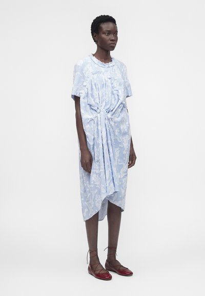 Γυναίκα που φοράει ένα ανοιχτό μπλε, υφασμάτινο φόρεμα μήκους μίντι με ραμμένες λεπτομέρειες στο μπροστινό μέρος και κόκκινα δερμάτινα παπούτσια με κορδόνια, στέκεται μπροστά σε λευκό φόντο.