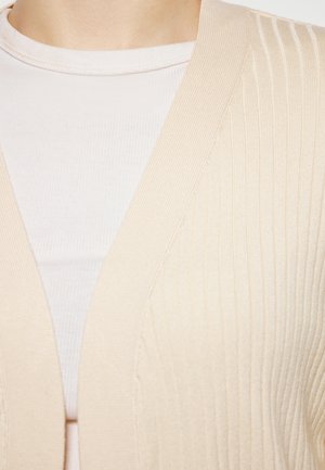 Beige geribde cardigan met een V-hals ontwerp. Zachte textuur met verticale strepen, gedragen over een lichtgekleurde, nauwsluitende top.