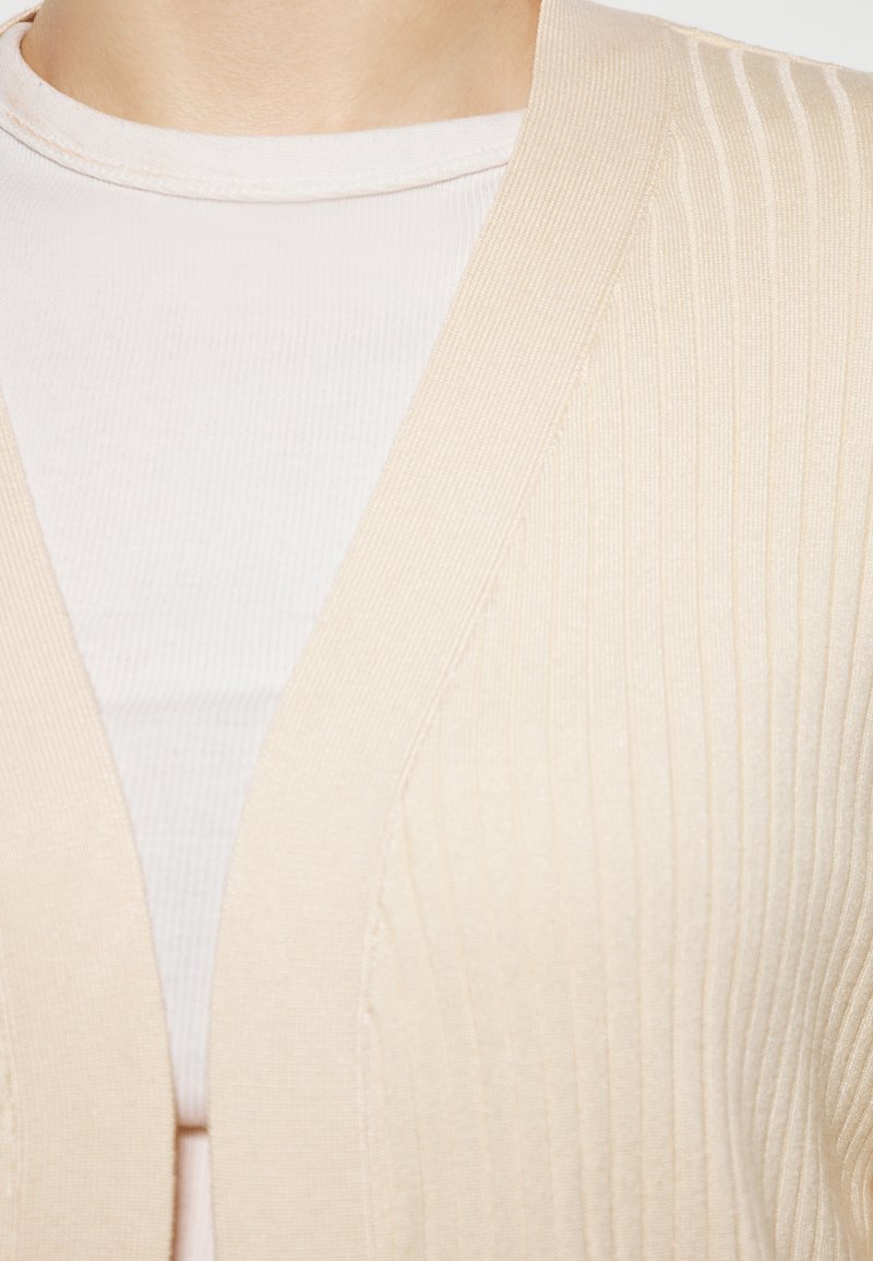 Cardigan beige a costine con scollatura a V. Texture morbida con strisce verticali, indossato sopra un top aderente di tonalità chiara.
