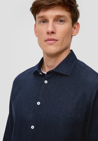 Granatowa lniana koszula z kołnierzykiem i białymi guzikami, wyróżniająca się teksturowanym materiałem i luźnym krojem. Minimalistyczny design, bez wzorów.