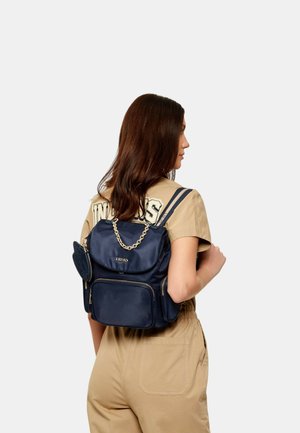 Donna con capelli castani lunghi che indossa una tuta beige e porta uno zaino Liu Jo blu navy con dettaglio a catena dorata, vista di lato.