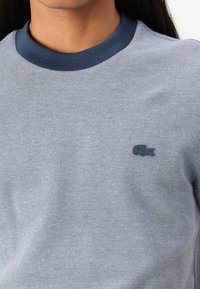 Sivý pletený svetric s námorníckym ribkovaným výstrihom a diskrétnym tmavomodrým vyšitým logom Lacoste na ľavej hrudi.