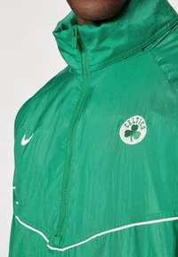 Férfi zöld, cipzáras Celtics sportdzsekit visel, fehér Nike logóval és csapatemblémával a mellkason.