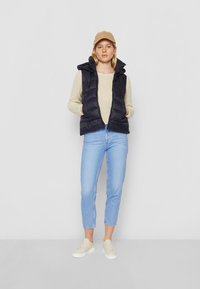 Kobieta w beżowej czapce, kremowym swetrze, granatowej kamizelce puchowej, jasnoniebieskich dżinsach i białych trampkach stoi na tle czystej białej ściany.