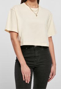 Donna che indossa una maglietta cropped a maniche corte beige, jeans neri a vita alta e collane d'oro a strati con un pendente a forma di cuore.
