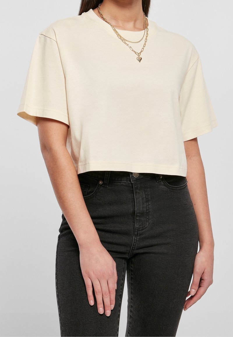 Donna che indossa una maglietta cropped a maniche corte beige, jeans neri a vita alta e collane d'oro a strati con un pendente a forma di cuore.