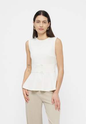 Vrouw met lang donker haar draagt een mouwloze witte peplum top met kleine strikjes in de taille en beige broek, staand tegen een effen achtergrond.
