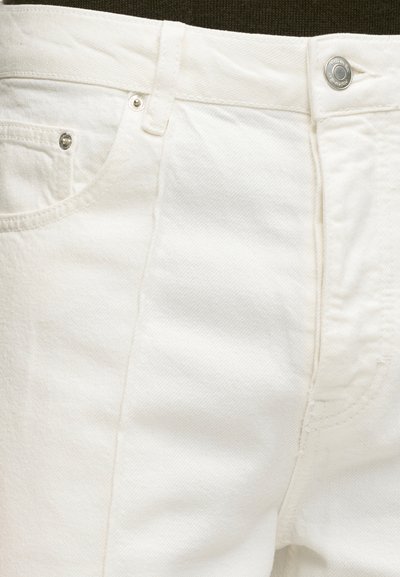 Gros plan sur un jean en denim blanc montrant la poche avant, la boucle de ceinture et le bouton métallique avec gravure de la marque.