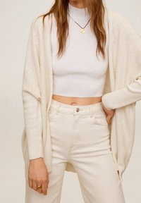 Cardigan en tricot de couleur crème avec un devant ouvert, des manches longues et une coupe ample, superposé à un crop top blanc en maille côtelée et à un jean beige taille haute.