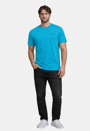 Mann steht mit der rechten Hand in der Hosentasche, trägt ein leuchtend blaues Kurzarm-T-Shirt, schwarze Jeans und weiße Sneakers vor einem einfarbigen Hintergrund.