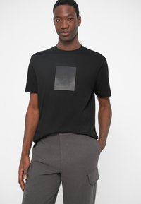 Μαύρο βαμβακερό t-shirt με τετράγωνο γραφικό σχέδιο σε αποχρώσεις του γκρι στο στήθος. Κοντά μανίκια και σχέδιο με στρογγυλή λαιμόκοψη.