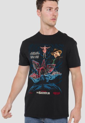 Man wearing black Stranger Things T-shirt with graphic of Demogorgon, characters, and text about an underground gate near a water tank.