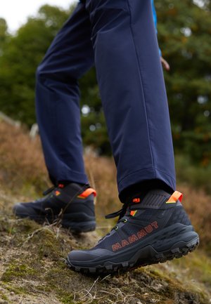 Schwarze Wanderstiefel mit einer strukturierten Überlage, leuchtenden orangefarbenen Akzenten und einer robusten Sohle, kombiniert mit marineblauen Outdoor-Hosen auf unebenem Gelände.