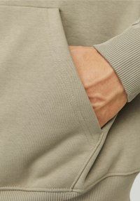 Sudadera beige con un bolsillo frontal. La tela es suave, con un puño de canal�é, que muestra detalles de costura sutiles y un corte relajado.