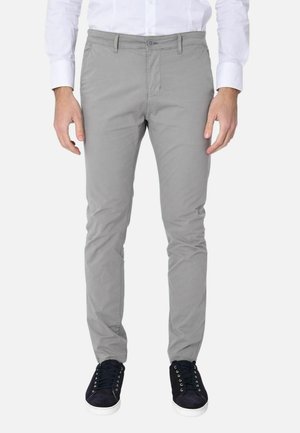 Uomo che indossa pantaloni grigio chiaro aderenti, camicia bianca elegante e sneaker nere con suole bianche, in piedi davanti a uno sfondo bianco.