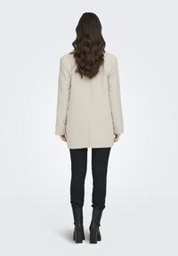 Blazer beige ajustado con corte recto, combinado con jeans negros ajustados y botas negras hasta la rodilla. Tejido suave y mangas largas.