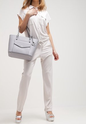 Femme en combinaison blanche et talons plateforme argentés tenant un sac cabas gris Michael Kors avec un porte-clés logo.