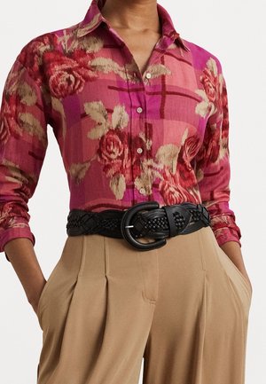 Kobieta w różowej koszuli z kwiatowym wzorem, zapinanej na guziki, włożonej w spodnie beżowe z wysokim stanem i plisami, z czarnym plecionym paskiem. Ręce w kieszeniach.