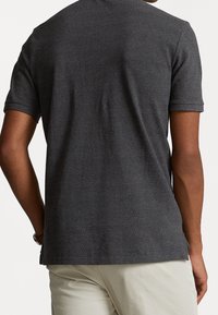 Mann in einem dunkelgrauen kurzärmeligen Poloshirt und hellbeigen Hosen, von hinten vor einem schlichten hellen Hintergrund gesehen.