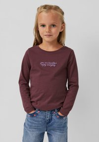 Langärmliges weinrotes Shirt mit dem Text "Du bist schön, genieße jeden Tag" in lila, kombiniert mit blauer Jeans, die florale Stickereien auf den Taschen aufweist.