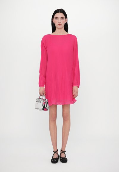 Roze geplooide jurk met lange mouwen en een vierkante halslijn, gecombineerd met een zilveren handtas en zwarte schoenen met een stippenpatroon.
