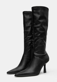 Fekete bőr bokacsizma hegyes orral, magas stiletto sarkal és cipzáras záródással. Simító felülettel és testhezálló dizájnnal rendelkezik.