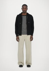 Felpa nera con zip, t-shirt a righe, pantaloni beige chiaro a gamba larga e scarpe nere. Design semplice con cappuccio regolabile e tasca.