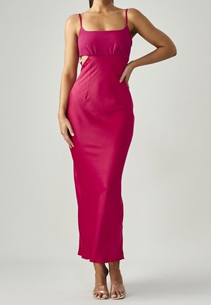 Kobieta w różowej, bezrękawnikowej sukni maxi z wycięciami na talii i przezroczystych sandałach na wysokim obcasie, stojąca na jednolitym tle.