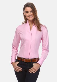 Camicia button-up rosa chiaro con maniche lunghe, colletto moderno e pieghe verticali. Abbinata a jeans scuri e una cintura marrone.