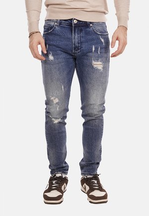 Jeans attillati in denim blu scuro con toppe strappate, texture sfilacciata, passanti per cintura e tasche frontali. Indossati con sneakers marroni e bianche.