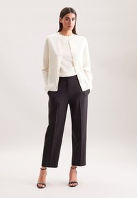 Weißer Cardigan über einem cremefarbenen Oberteil, kombiniert mit schwarzen Hosen. Verfügt über eine Knopfleiste, einen lockeren Schnitt und schwarze Ankle-Strap-Absätze.