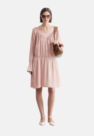 Kobieta ubrana w jasnoróżową sukienkę z długim rękawem, duże okulary przeciwsłoneczne, białe baleriny i niosąca brązową torebkę na ramię na jednolitym tle.