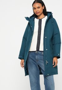 Casaco de inverno longo em tom teal, com capuz, fecho de zip e botões de pressão, usado sobre um suéter texturizado em creme e jeans azuis.