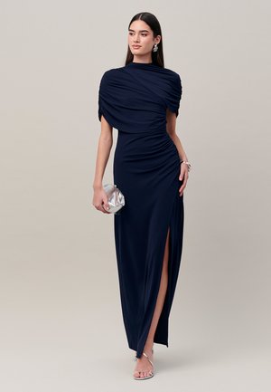 Vrouw in een marineblauwe avondjurk met geplooid detail en een hoge split, houdt een zilveren clutch vast, draagt zilveren hakken en oorbellen, staand tegen een effen achtergrond.