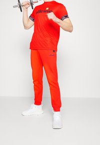 Rød atletisk outfit med en t-shirt med logo og mønstrede ærmer, parret med matchende sweats og hvide sneakers. Vægt i hånden.