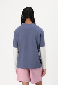 T-skjorte med korte ermer i blå bomull med en lys grå langermet skjorte under, brukt med rosa shorts, som viser en avslappet passform og enkel design.