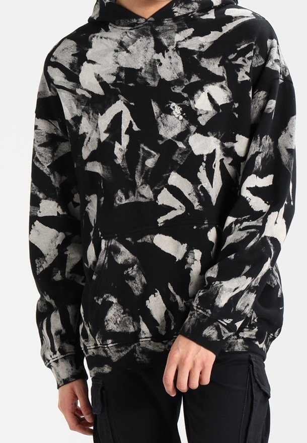 Sweat à capuche noir avec des motifs abstraits de coups de pinceau blancs et gris porté par une personne avec les mains détendues le long du corps.