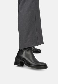 Botas de tobillo de cuero negro con punta redonda, tacón bajo y textura suave; combinadas con pantalones de sastre grises.