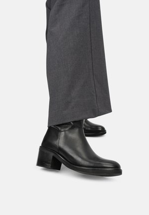 Bottines en cuir noir avec un bout rond, un talon bas et une texture lisse ; associées à un pantalon gris ajusté.
